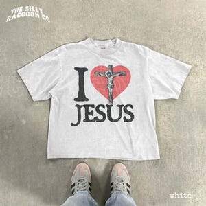 A white t-shirt with a graphic design of a cross and the text "I ♥ JESUS" printed on it, along with a heart symbol. The t-shirt is laid out on a concrete floor, and a pair of white sneakers is visible underneath it.