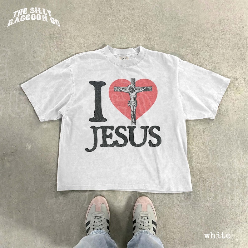 A white t-shirt with a graphic design of a cross and the text "I ♥ JESUS" printed on it, along with a heart symbol. The t-shirt is laid out on a concrete floor, and a pair of white sneakers is visible underneath it.