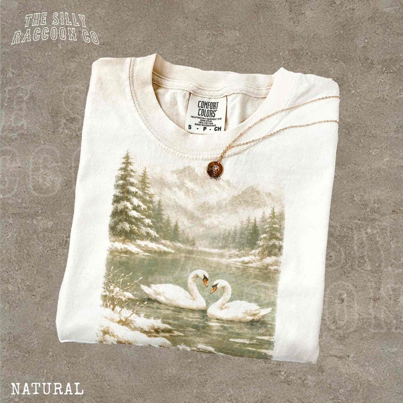 A white t-shirt with a printed image of two swans swimming in a lake, surrounded by snow-covered trees.