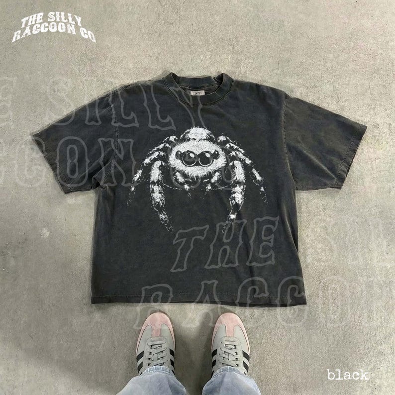 A pair of black sneakers and a black t-shirt with a spider graphic printed on it, laid out on a concrete floor.