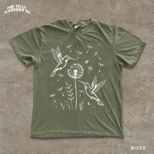 A green t-shirt with a white illustration of two hummingbirds hovering over a dandelion surrounded by flowers and plants.