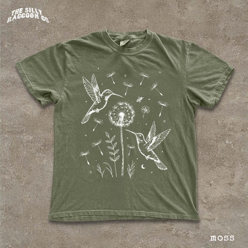 A green t-shirt with a white illustration of two hummingbirds hovering over a dandelion surrounded by flowers and plants.