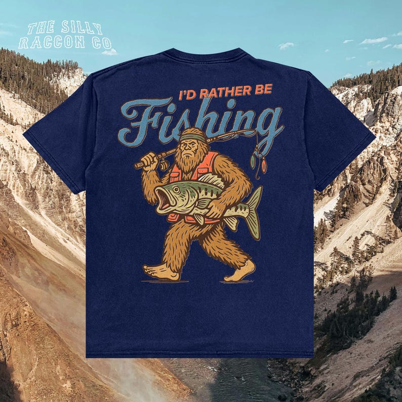 A blue t-shirt with a graphic design depicting a large, furry creature holding a fish and the text "I'D Rather Be Fishing" printed on it.