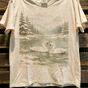 A white t-shirt with a printed image of two swans swimming in a lake, surrounded by snow-covered trees.