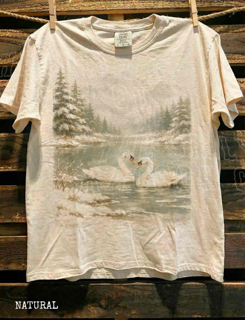 A white t-shirt with a printed image of two swans swimming in a lake, surrounded by snow-covered trees.