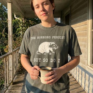 A person wearing a gray t-shirt with a graphic of a rat and the text "The Horrors Persist BUT SO DO I" is holding a mug and standing on a porch.
