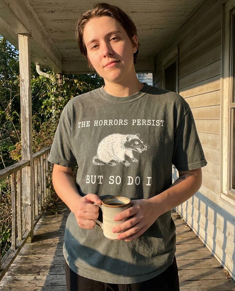 A person wearing a gray t-shirt with a graphic of a rat and the text "The Horrors Persist BUT SO DO I" is holding a mug and standing on a porch.