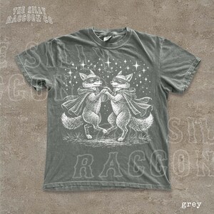 A grey t-shirt with a graphic design featuring two foxes dancing under a starry sky.