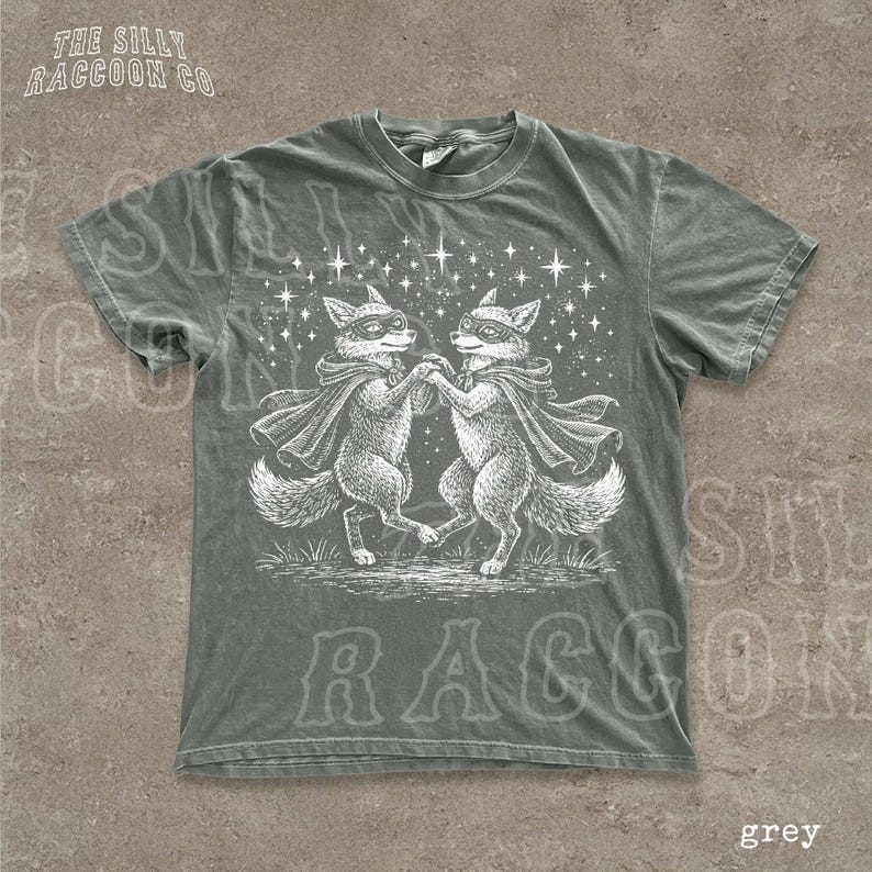 A grey t-shirt with a graphic design featuring two foxes dancing under a starry sky.