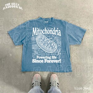 A blue t-shirt with a graphic design and text, along with a pair of sneakers, is displayed on a concrete floor.