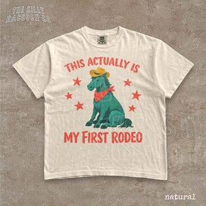a white t-shirt with a graphic design depicting a green horse wearing a cowboy hat and a red bandana, surrounded by red stars. The text on the t-shirt reads "This actually is my first rodeo" and is located in the center of the design.