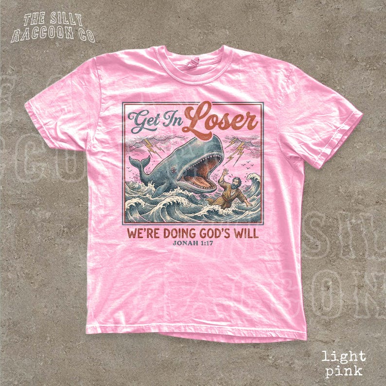 A pink t-shirt with a graphic design depicting a man being attacked by a large whale, with the text "Get In Loser" and "We're Doing God's Will" below the image.
