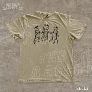 A beige t-shirt with a graphic design featuring three cats, each with a unique pose, and the text "THE SILKY RACCOON CO." printed on it.