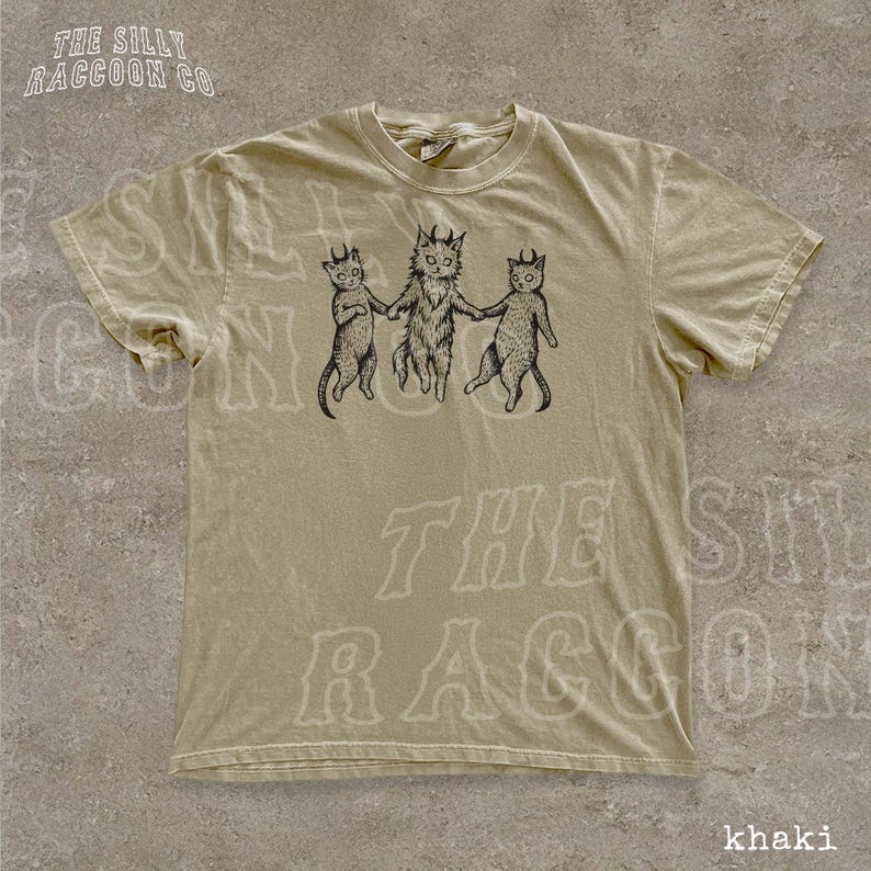 A beige t-shirt with a graphic design featuring three cats, each with a unique pose, and the text "THE SILKY RACCOON CO." printed on it.