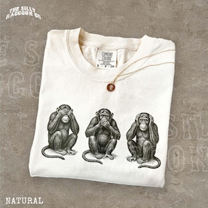 Op de afbeelding: Een natuurkleurig t-shirt met een zwarte inktillustratie van de drie wijze apen, waarbij één aap de ogen bedekt, één de mond en één de oren. Het shirt heeft een ronde hals en een Comfort Colors merklabel aan de binnenkant van de kraag.