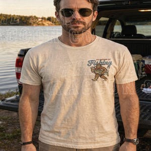 A man wearing sunglasses and a t-shirt with a fishing graphic stands in front of a pickup truck near a body of water.