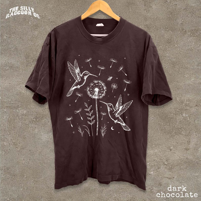 A dark brown t-shirt with a white illustration of two hummingbirds and a dandelion on a branch, along with other floral and natural elements.