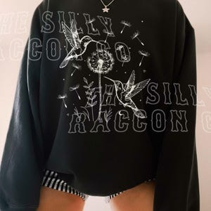 A person wearing a black sweatshirt with a graphic design featuring a bird and a dandelion.