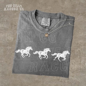 a gray t-shirt with a graphic design of three white horses running across the front. The t-shirt is displayed on a textured surface, and there is a small necklace visible around the neck of the shirt.