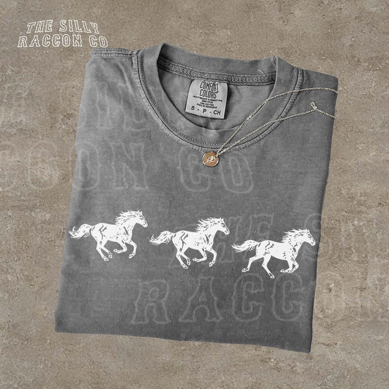 a gray t-shirt with a graphic design of three white horses running across the front. The t-shirt is displayed on a textured surface, and there is a small necklace visible around the neck of the shirt.