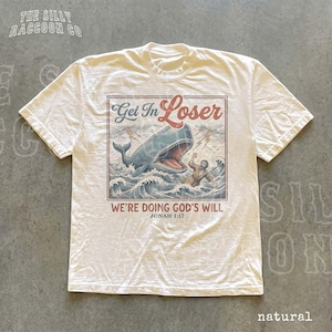 A white t-shirt with a graphic design depicting a person being attacked by a large whale, with the text "Get In Loser" and "We're Doing God's Will" printed on it.