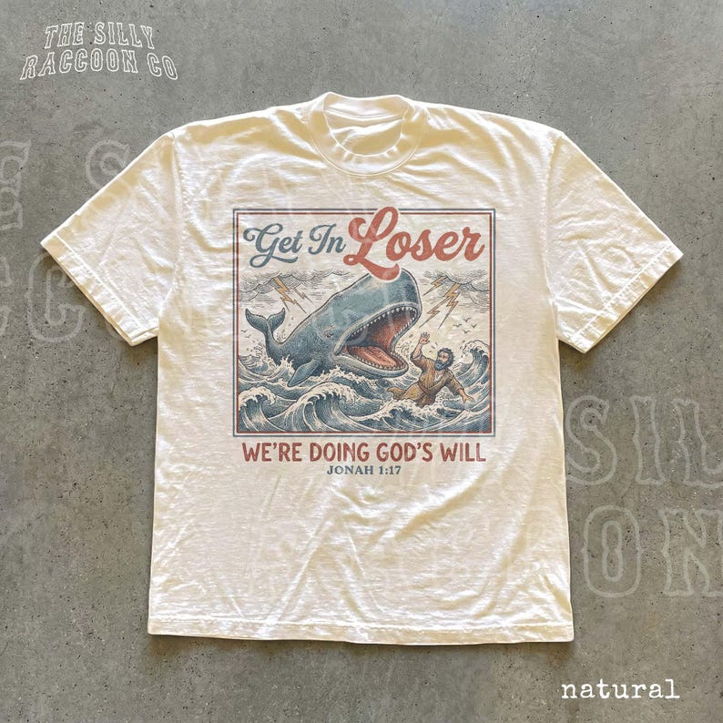 A white t-shirt with a graphic design depicting a person being attacked by a large whale, with the text "Get In Loser" and "We're Doing God's Will" printed on it.