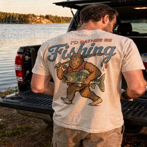 A man standing in front of a truck with a t-shirt featuring a cartoon fish and the text "I'D Rather Be Fishing".