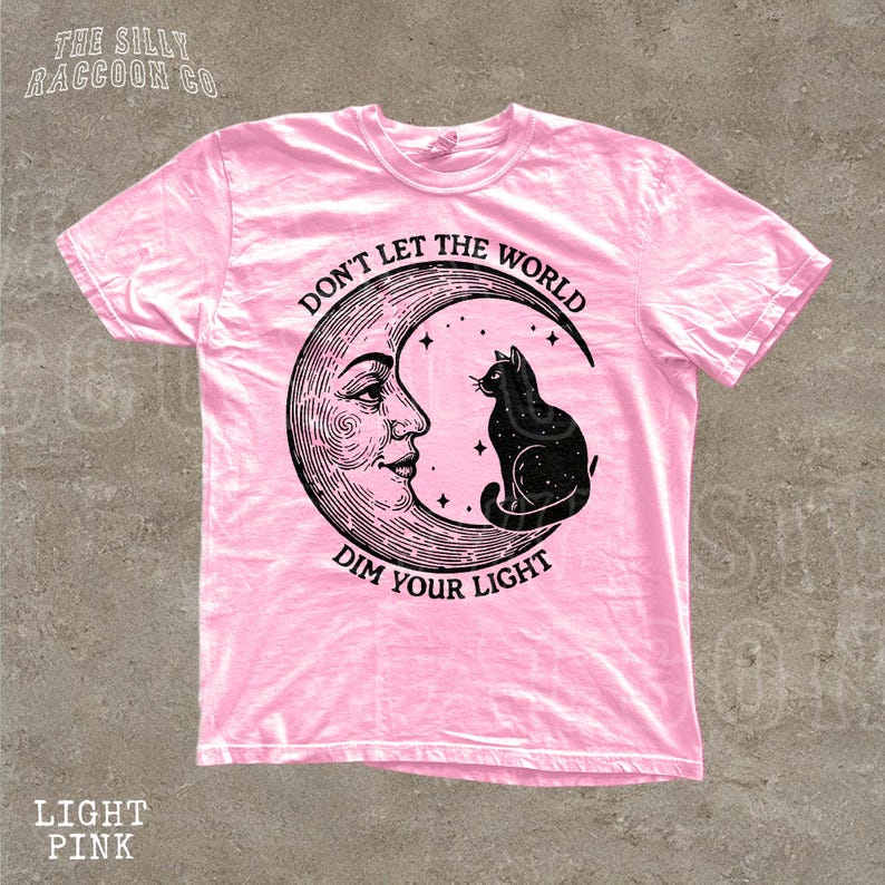 A pink t-shirt with a graphic design featuring a cat sitting on a crescent moon and the text "Don't let the world dim your light" printed on it.