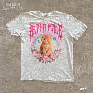 A white t-shirt with a graphic design featuring an orange cat surrounded by a rainbow, flowers, and butterflies.