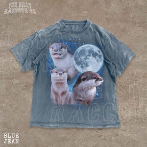 A t-shirt with a graphic design featuring three otters against a backdrop of a full moon and stars.