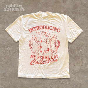 A white t-shirt with a graphic design featuring three cats and the text "INTRODUCING MY FERAL CAT COLLECTION" printed on it.