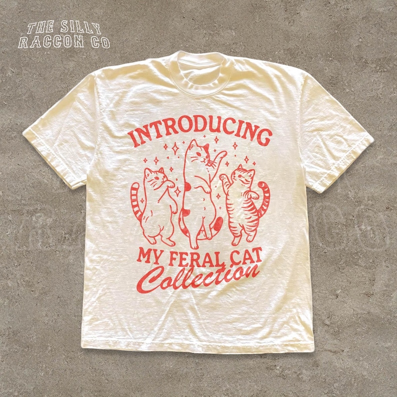 A white t-shirt with a graphic design featuring three cats and the text "INTRODUCING MY FERAL CAT COLLECTION" printed on it.