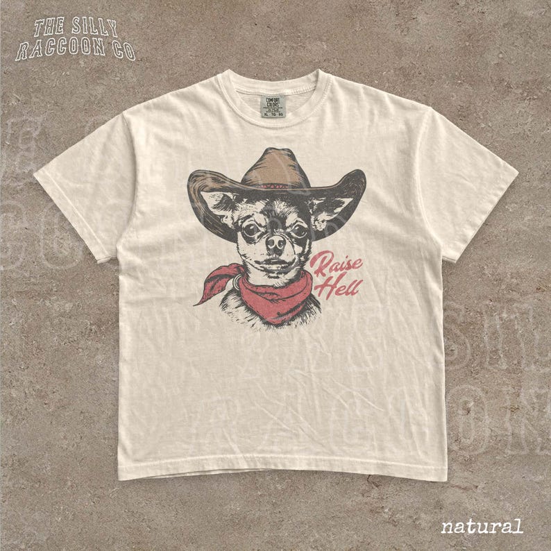 A white t-shirt with a graphic design of a dog wearing a cowboy hat and a red scarf, along with the text "Raise Hell" printed on it.