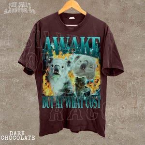 A dark brown t-shirt with a graphic design featuring two koalas and the text "AWAKE BUT AT WHAT COST" printed on it.