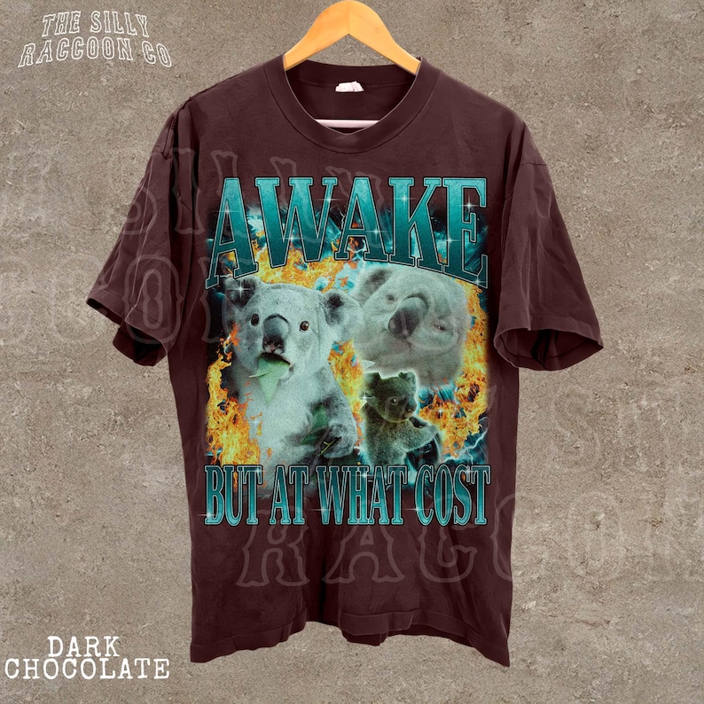 A dark brown t-shirt with a graphic design featuring two koalas and the text "AWAKE BUT AT WHAT COST" printed on it.