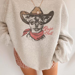 A person wearing a gray sweatshirt with a graphic of a dog wearing a cowboy hat and a red scarf, along with the text "Raise Hell" printed on it.