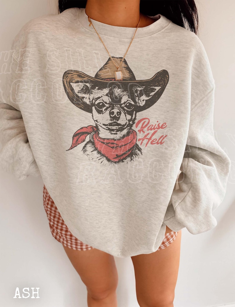 A person wearing a gray sweatshirt with a graphic of a dog wearing a cowboy hat and a red scarf, along with the text "Raise Hell" printed on it.