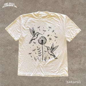 A white t-shirt with a black and white graphic design depicting two hummingbirds and a dandelion surrounded by flowers and leaves.