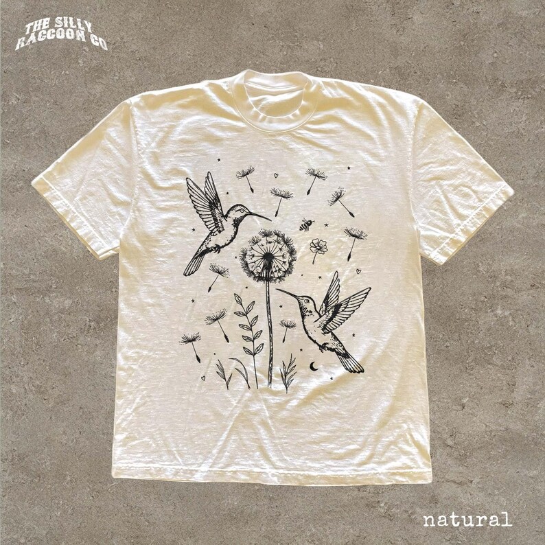 A white t-shirt with a black and white graphic design depicting two hummingbirds and a dandelion surrounded by flowers and leaves.
