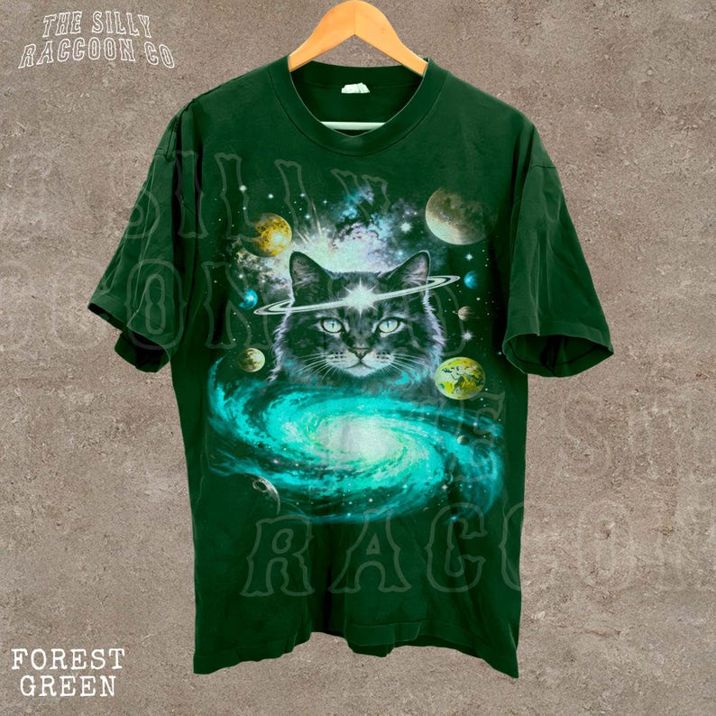 A green t-shirt with a cat wearing a helmet and surrounded by planets and a swirling galaxy design.