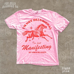 A pink t-shirt with a graphic of a running horse and text that reads "I'm not delusional, I'm just Manifesting my own reality".