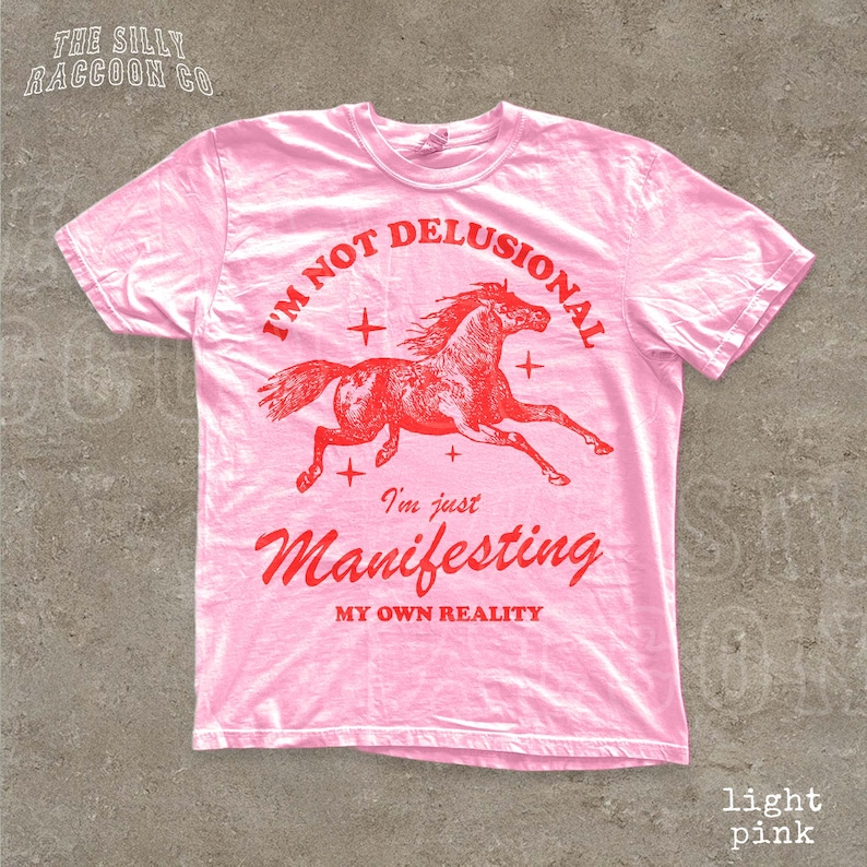 A pink t-shirt with a graphic of a running horse and text that reads "I'm not delusional, I'm just Manifesting my own reality".
