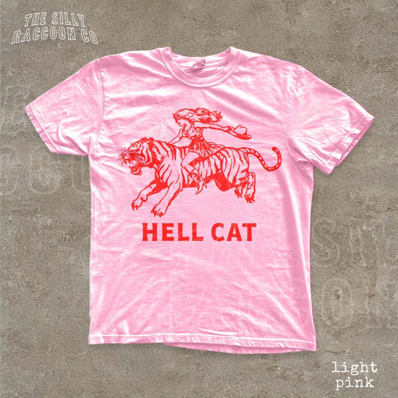 A pink t-shirt with a graphic design depicting a person riding a tiger, accompanied by the text "HELL CAT" in red letters.