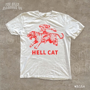 A white t-shirt with a red graphic design depicting a person riding a tiger. The text below the graphic says "HELL CAT".