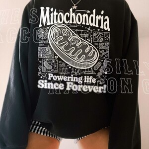 a person wearing a black sweatshirt with a graphic design depicting Mitochondria, a cellular organelle, and text that reads "Mitochondria" and "Powering life since forever!".