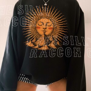A person wearing a black sweatshirt with a sun design and mountains printed on it.