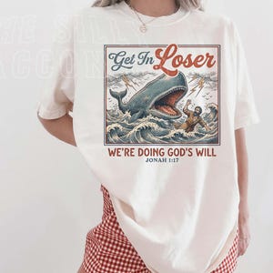 A woman wearing a white t-shirt with a graphic design depicting a person being attacked by a large whale, along with the text "Get In Loser" and "We're Doing God's Will" below the image.