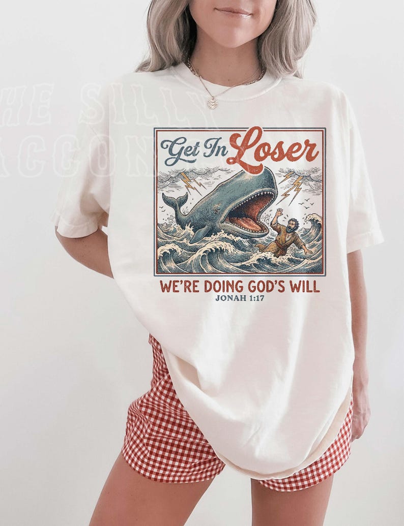 A woman wearing a white t-shirt with a graphic design depicting a person being attacked by a large whale, along with the text "Get In Loser" and "We're Doing God's Will" below the image.