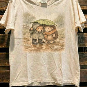 A t-shirt with a printed illustration of two hedgehog characters standing together under an umbrella, with a wooden background.
