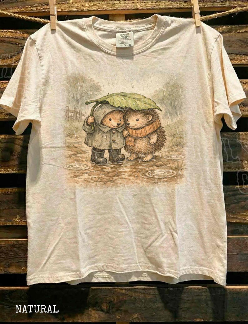 A t-shirt with a printed illustration of two hedgehog characters standing together under an umbrella, with a wooden background.